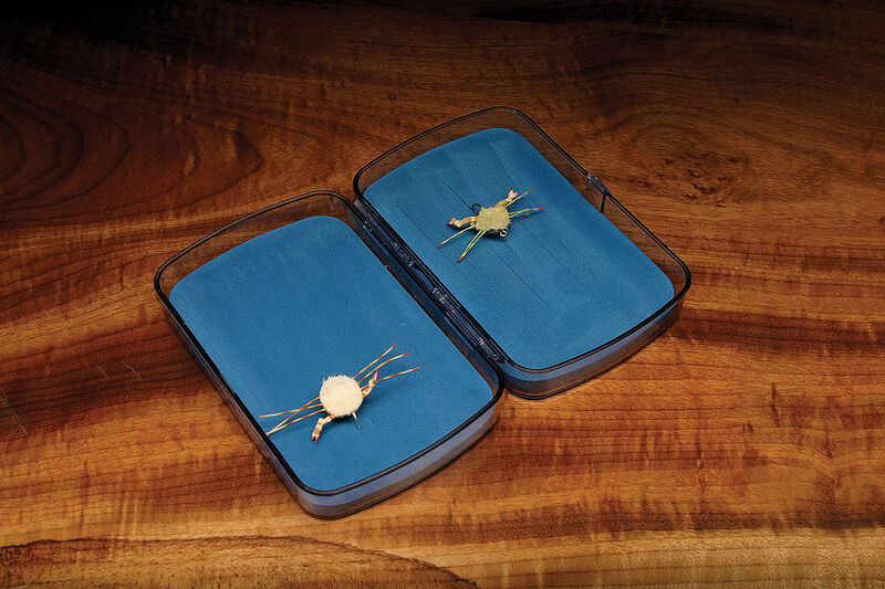 Open fly box with blue interior holding two crab fly patterns on a wooden table.