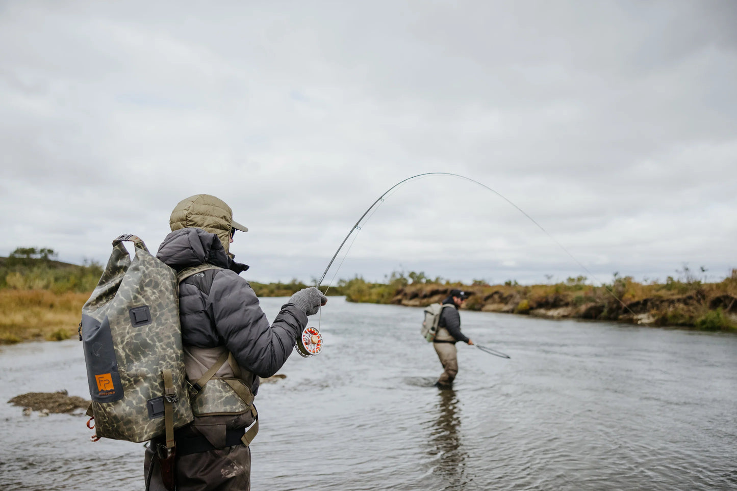 Angler standing in a river while fly fishing, wearing the Fishpond Wind River Roll-Top Backpack.