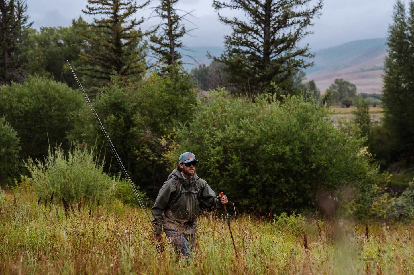 Angler walking through tall grass with a bamboo Fishpond wading staff and fly fishing gear