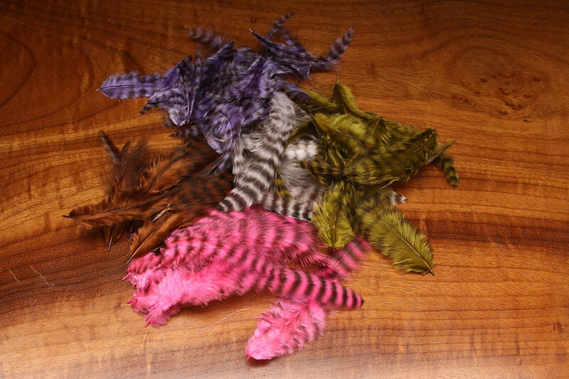 Assorted UV2 Grizzly Soft Hackle feathers in multiple colors including pink, olive, brown, purple, and natural, displayed on a wood surface.